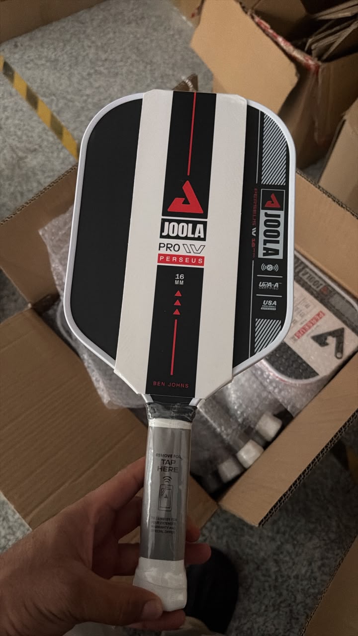 A hand holding a Joola Pro W11 pickleball paddle in its protective plastic wrapping, with cardboard boxes in the background.