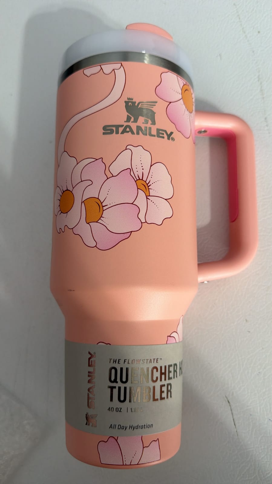 A pink Stanley Quenchers Tumbler with a floral pattern, a matching pink handle, and a gray label at the bottom, photographed on a light gray surface.