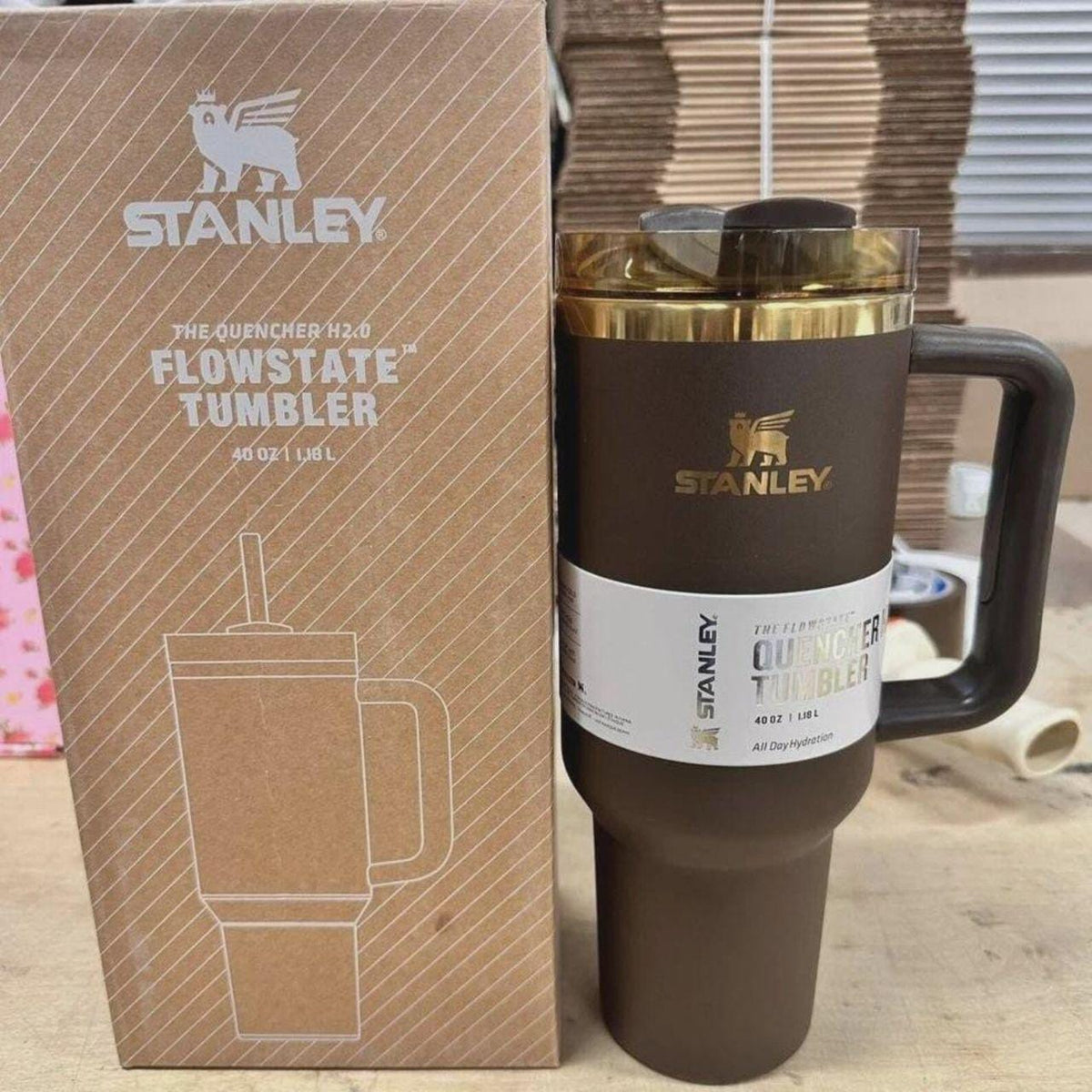 A dark brown Stanley Quencher 40 oz tumbler with a gold lid trim and a white label, standing next to its brown cardboard packaging box on a wooden surface.