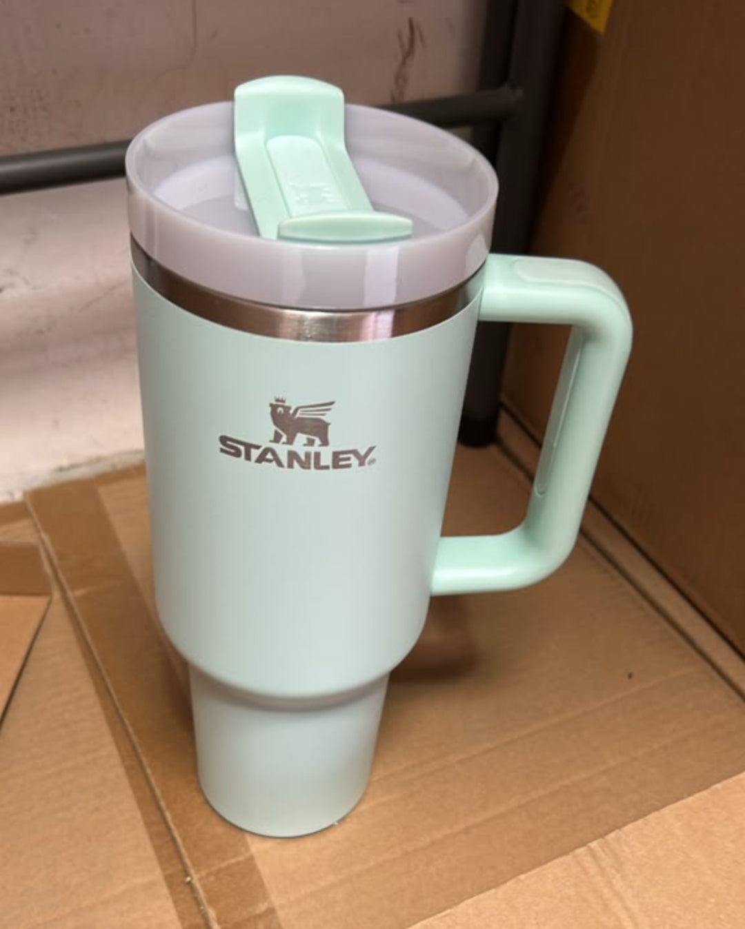 A mint green Stanley travel mug with a matching lid and handle, resting on a cardboard box.