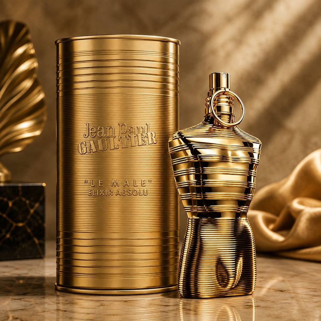 Jean Paul Gaultier Le Male Lux Elixir Absolu perfume bottle and matching cylindrical packaging, both in a polished gold finish, displayed on a marble surface with draped fabric in the background.