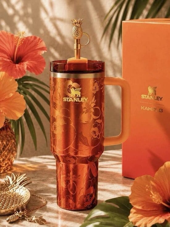 A Stanley Kahito 16 oz tumbler in a metallic copper-orange finish with a floral pattern, shown next to its orange packaging box. The tumbler features a matching handle and a gold-toned straw with a pineapple topper, styled with tropical flowers and leaves.