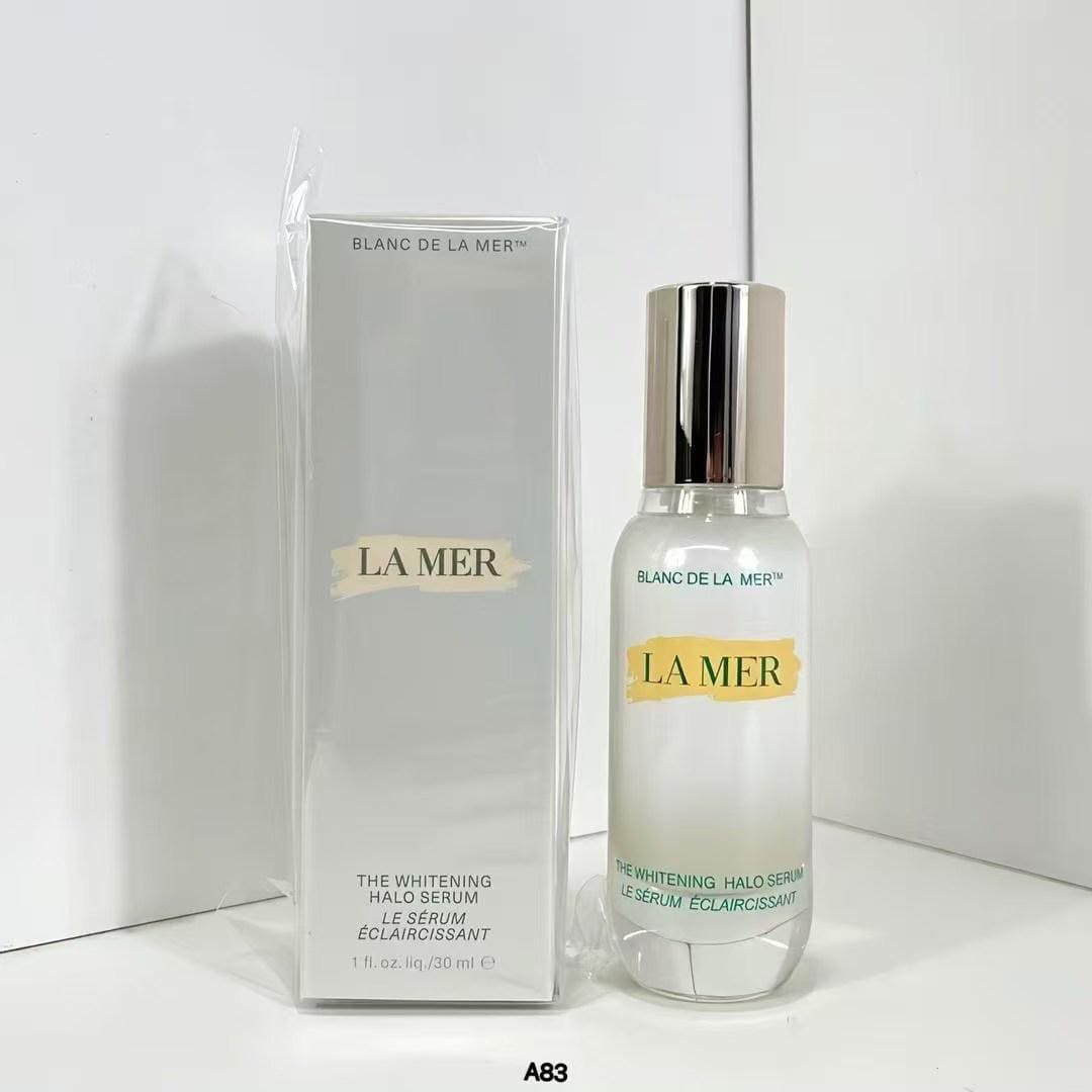 La Mer The Whitening Halo Serum shown next to its original packaging box. The clear bottle features a metallic silver cap and a gold label with the brand name 'LA MER'.