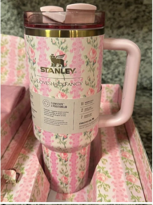 A pink Stanley travel tumbler with a floral pattern, shown inside its original packaging box. The tumbler features a light pink handle, a clear lid with a pink flip-straw, and a gold band near the rim.
