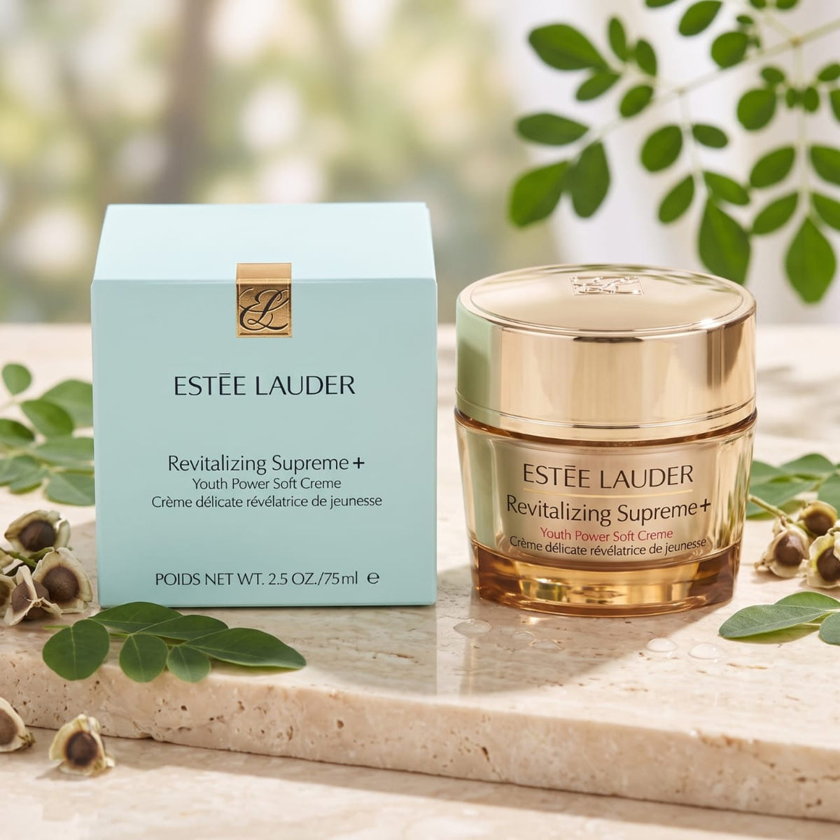 Estée Lauder Revitalizing Supreme+ Youth Power Soft Creme in a gold jar next to its light blue box, styled on a marble surface with green leaves.