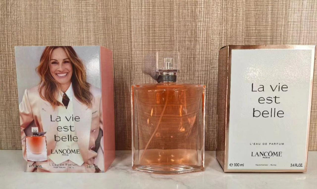 Lancôme La Vie Est Belle Eau de Parfum 100ml bottle with its pink and white outer boxes, displayed on a light marble surface against a textured beige wall.