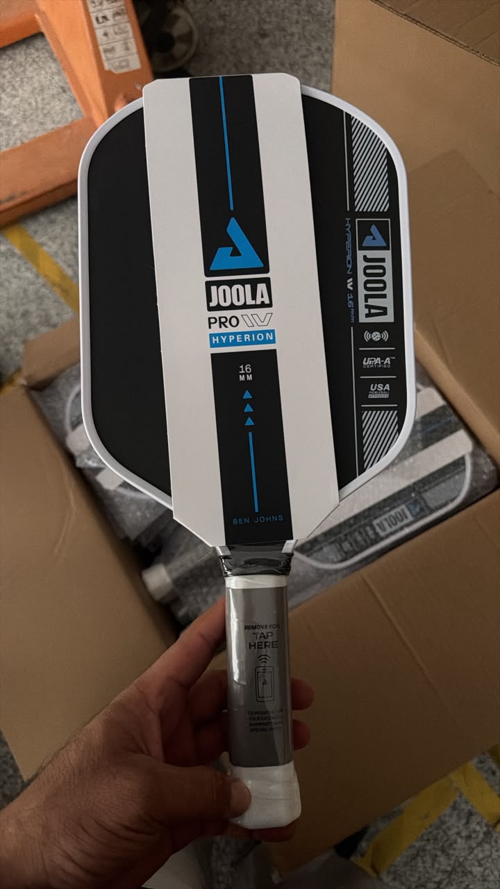 A hand holding a Joola Pro Hyperion paddle in a warehouse or shipping area, with cardboard boxes visible in the background.
