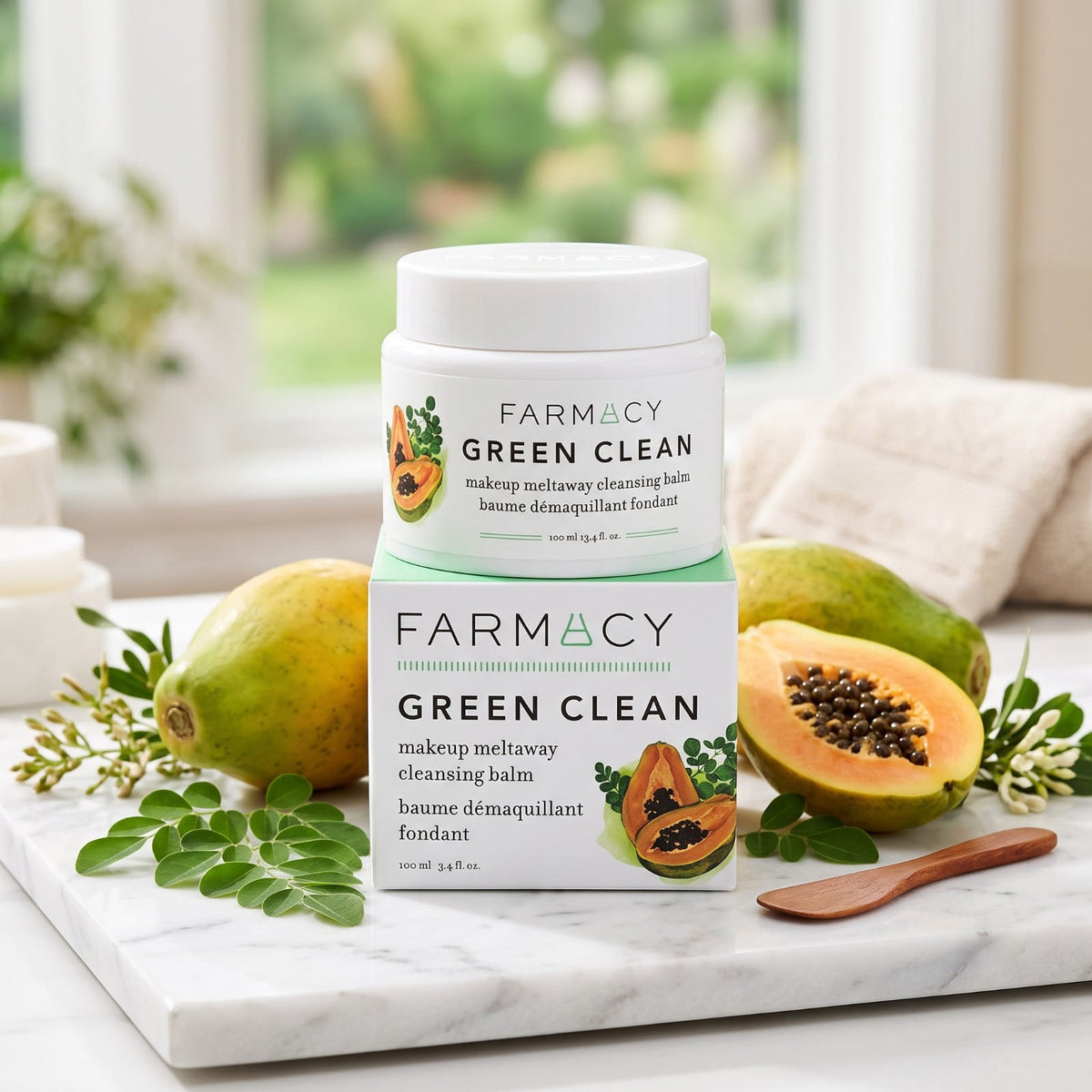 A jar of FARMACY GREEN CLEAN makeup meltaway cleansing balm is stacked on top of another jar and displayed on a marble tray with papayas, green leaves, and a wooden spoon in a bright, natural setting.