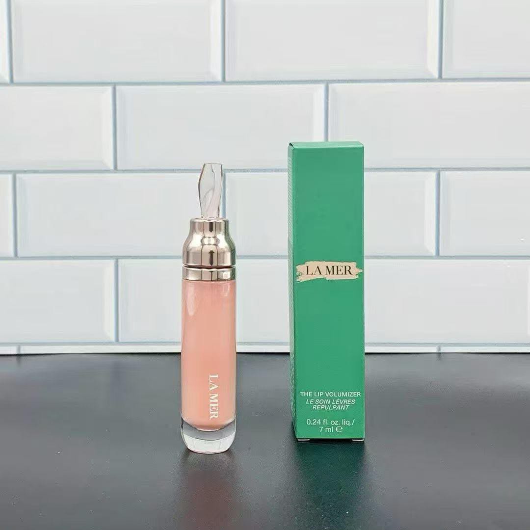 La Mer The Lip Volumizer in a clear glass bottle with a metallic cap, shown next to its green packaging box on a dark surface against a white subway tile background.