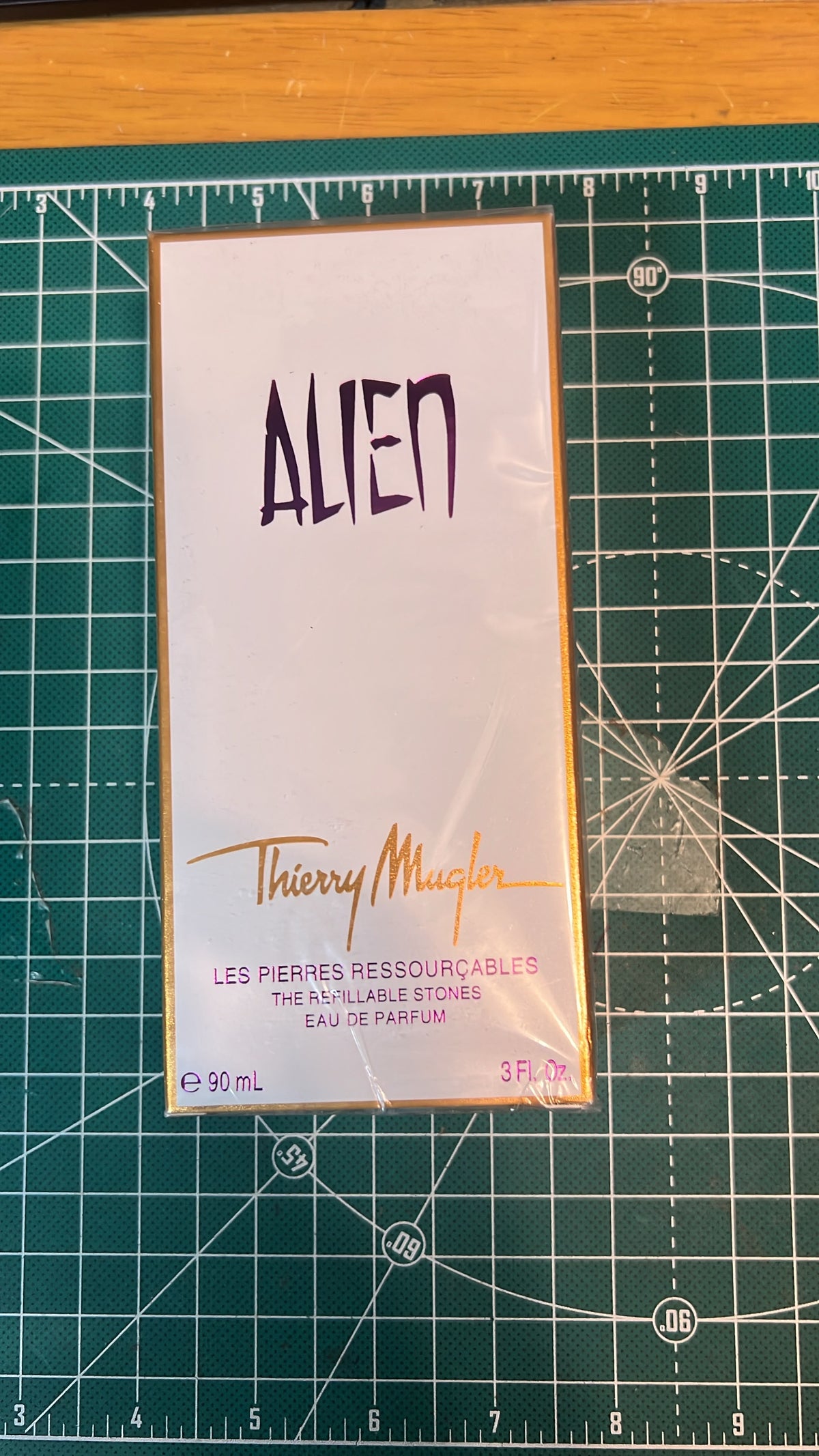 A 50 mL box of Thierry Mugler Alien perfume, still in its plastic shrink wrap, placed on a green cutting mat with a wooden table visible in the background.
