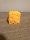 A small yellow cheese-shaped object with large holes, standing on a wooden surface against a plain light background.