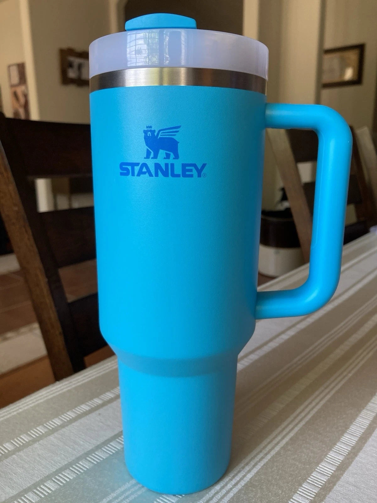 A bright blue Stanley travel mug with a handle and clear lid, standing on a striped tablecloth in a home setting.